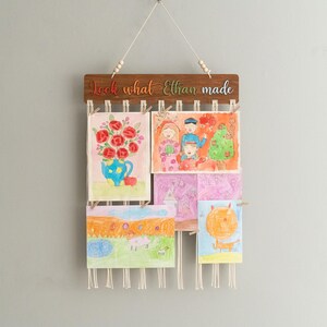 Puede incluir: Un colgador de pared de madera con el texto "Look what Ethan made" y una hilera de dibujos coloridos sujetos con pinzas de la ropa.