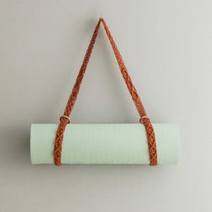 May include: A rolled-up, light green yoga mat suspended by a woven, rust-colored strap. The strap is looped through wooden rings and hangs from a single point, creating a triangular shape. The mat is against a plain, light-colored wall.