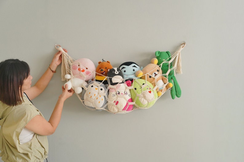 Op de afbeelding: Een witte macram&eacute; hangmat met een franje die aan een muur hangt, gevuld met verschillende knuffels, waaronder een groene kikker, een bruine beer, een roze varken, een witte uil en een zwart-witte skunk.