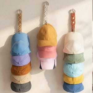 Peut inclure: Plusieurs casquettes de baseball de différentes couleurs, dont le bleu, le jaune, le rose et le vert, sont suspendues à un mur. Chaque casquette est empilée et suspendue par une sangle décorative en macramé. Les casquettes sont disposées verticalement, présentant un éventail de couleurs.