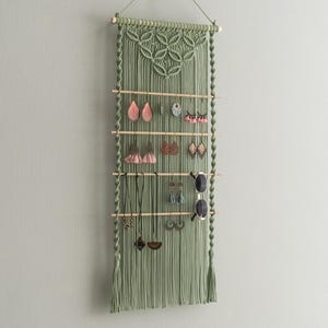 以下が含まれることがあります： 木製の棒とタッセルが付いた緑色のマクラメの壁掛け。ハンガーには、イヤリング、ネックレス、サングラスをかけるためのフックが複数列あります。