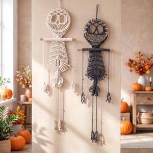 May include: Two macrame wall hangings shaped like skeletons, one in off-white and one in black. Each piece features a circular head, a ribcage design, and long dangling limbs with tassels, all suspended from a horizontal wooden dowel rod.