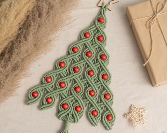 May include: A sage green macramé wall hanging shaped like a Christmas tree, featuring a wooden star at the top and red wooden beads woven throughout the cord. The piece ends in a tassel at the bottom, showcasing intricate knot work.