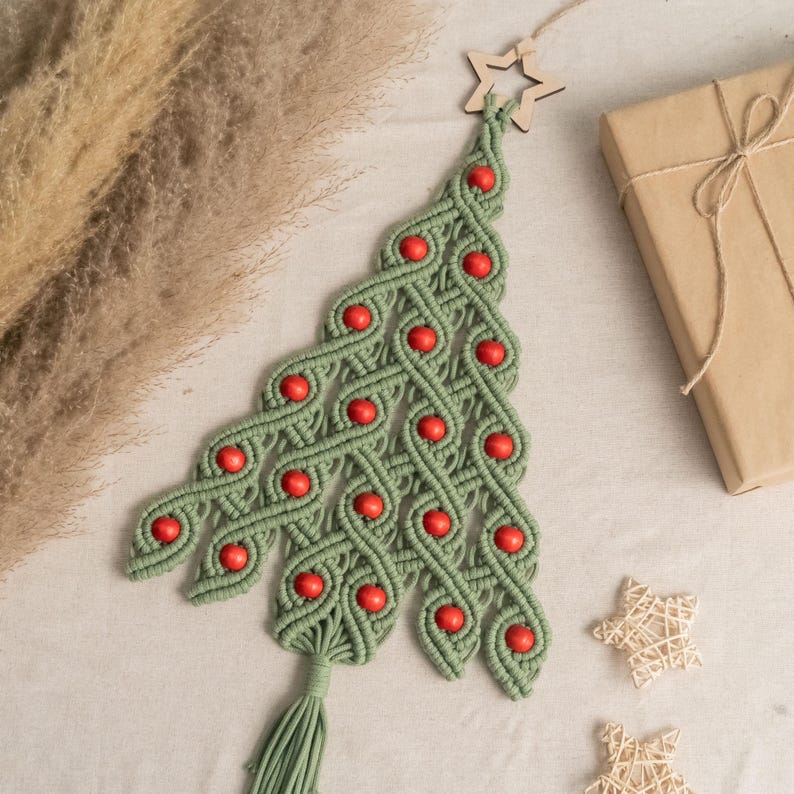 K&ouml;nnte beinhalten: Ein gr&uuml;ner Makramee-Weihnachtsbaum-Wandbehang mit roten Holzperlen. Der Baum ist mit einem Holzstern an der Spitze und einem Quasten an der Unterseite verziert.
