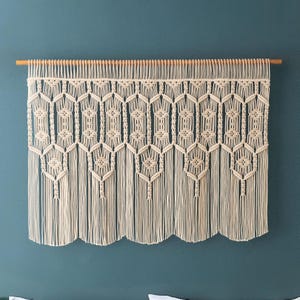 May include: A macrame wall hanging with a geometric pattern, made of natural cotton cord and hung on a wooden dowel rod. The wall hanging is cream colored and has a fringed bottom.
