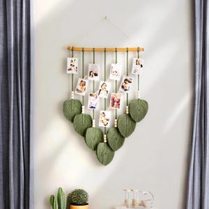 Pode incluir: Uma tape&ccedil;aria de macram&eacute; verde com uma barra de madeira e corda para pendurar fotos. A tape&ccedil;aria de parede &eacute; decorada com folhas verdes e contas de madeira.