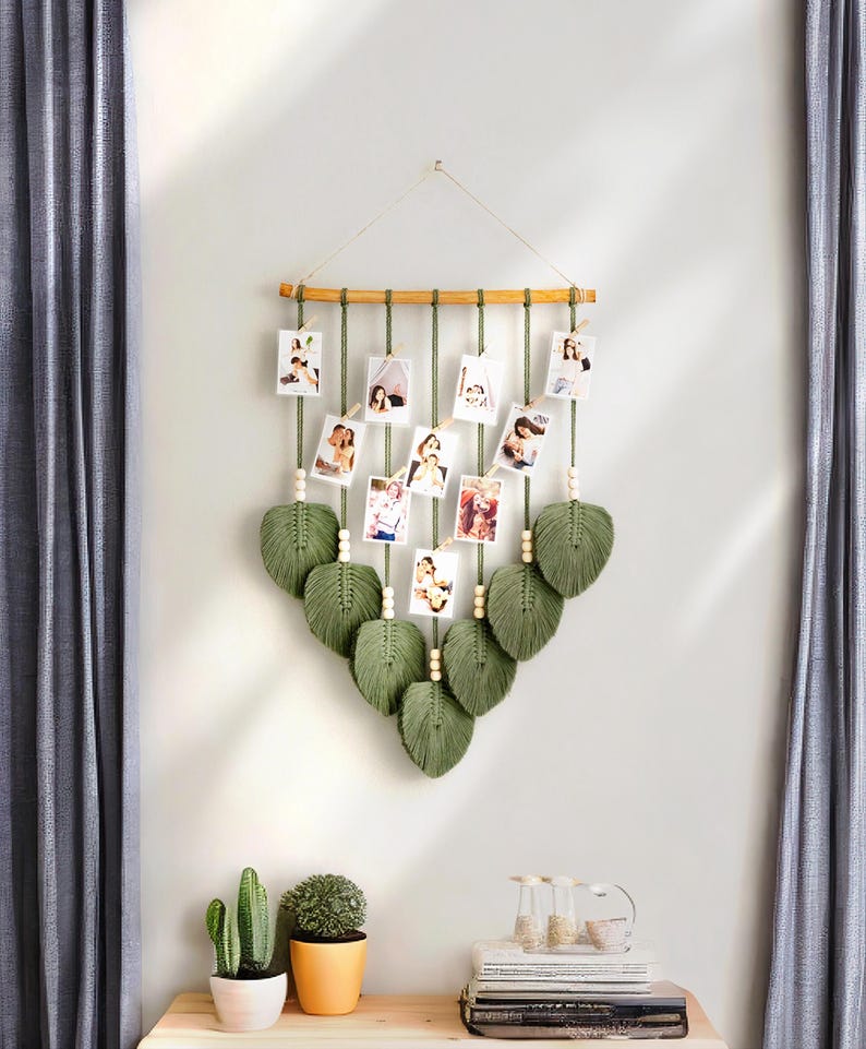 Pode incluir: Uma tape&ccedil;aria de macram&eacute; verde com uma barra de madeira e corda para pendurar fotos. A tape&ccedil;aria de parede &eacute; decorada com folhas verdes e contas de madeira.
