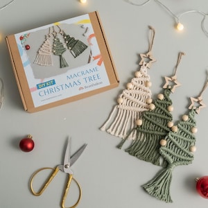 May include: A DIY macrame Christmas tree kit in a cardboard box labeled Macrame Christmas Tree BeanDaikon, featuring three finished macrame tree ornaments in cream and green with wooden beads and star toppers, plus gold-handled scissors.