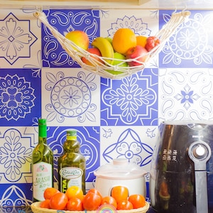 May include: A kitchen counter with a blue and white tiled backsplash. There are various items on the counter, including a white ceramic slow cooker, a black air fryer, two glass bottles of oil, a basket of red tomatoes, and a white string hammock filled with fruit.