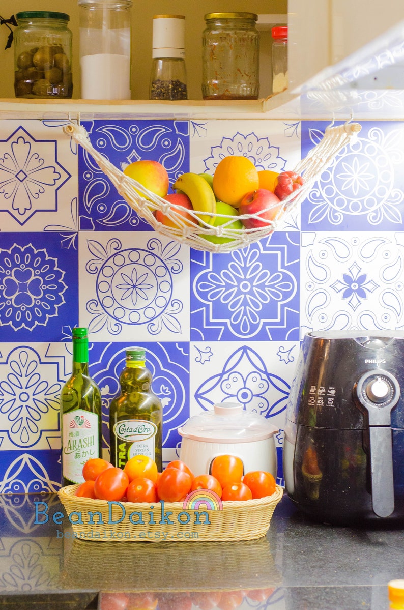 May include: A kitchen counter with a blue and white tiled backsplash. There are various items on the counter, including a white ceramic slow cooker, a black air fryer, two glass bottles of oil, a basket of red tomatoes, and a white string hammock filled with fruit.
