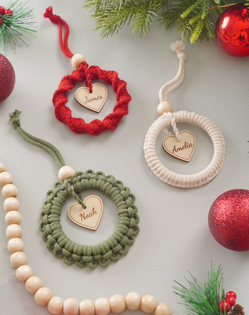 K&ouml;nnte beinhalten: Drei handgefertigte Weihnachtsornamente in Form von Kr&auml;nzen mit Holzh Herzen in der Mitte. Die Kr&auml;nze sind rot, wei&szlig; und gr&uuml;n. Die Herzen tragen die Namen "James", "Amelia" und "Noah".