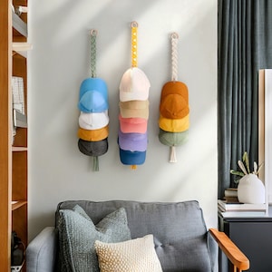 May include: Three wall-mounted hat organizers, each holding a stack of baseball caps in various colors. The organizers feature macrame or woven hanging straps with wooden rings. The caps include shades of blue, white, yellow, pink, and brown.