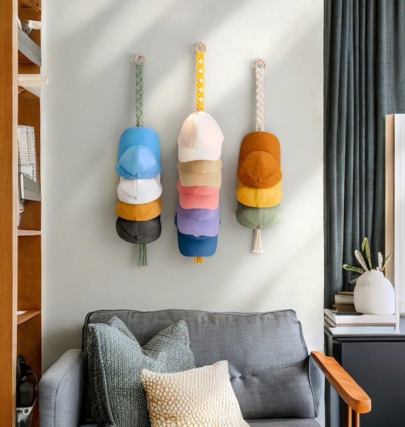 May include: Three wall-mounted hat organizers, each holding a stack of baseball caps in various colors. The organizers feature macrame or woven hanging straps with wooden rings. The caps include shades of blue, white, yellow, pink, and brown.
