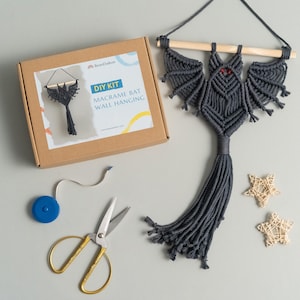 May include: DIY kit for making a macrame bat wall hanging. The kit includes grey yarn, a wooden dowel, a measuring tape, and scissors. The finished bat is grey with red eyes.