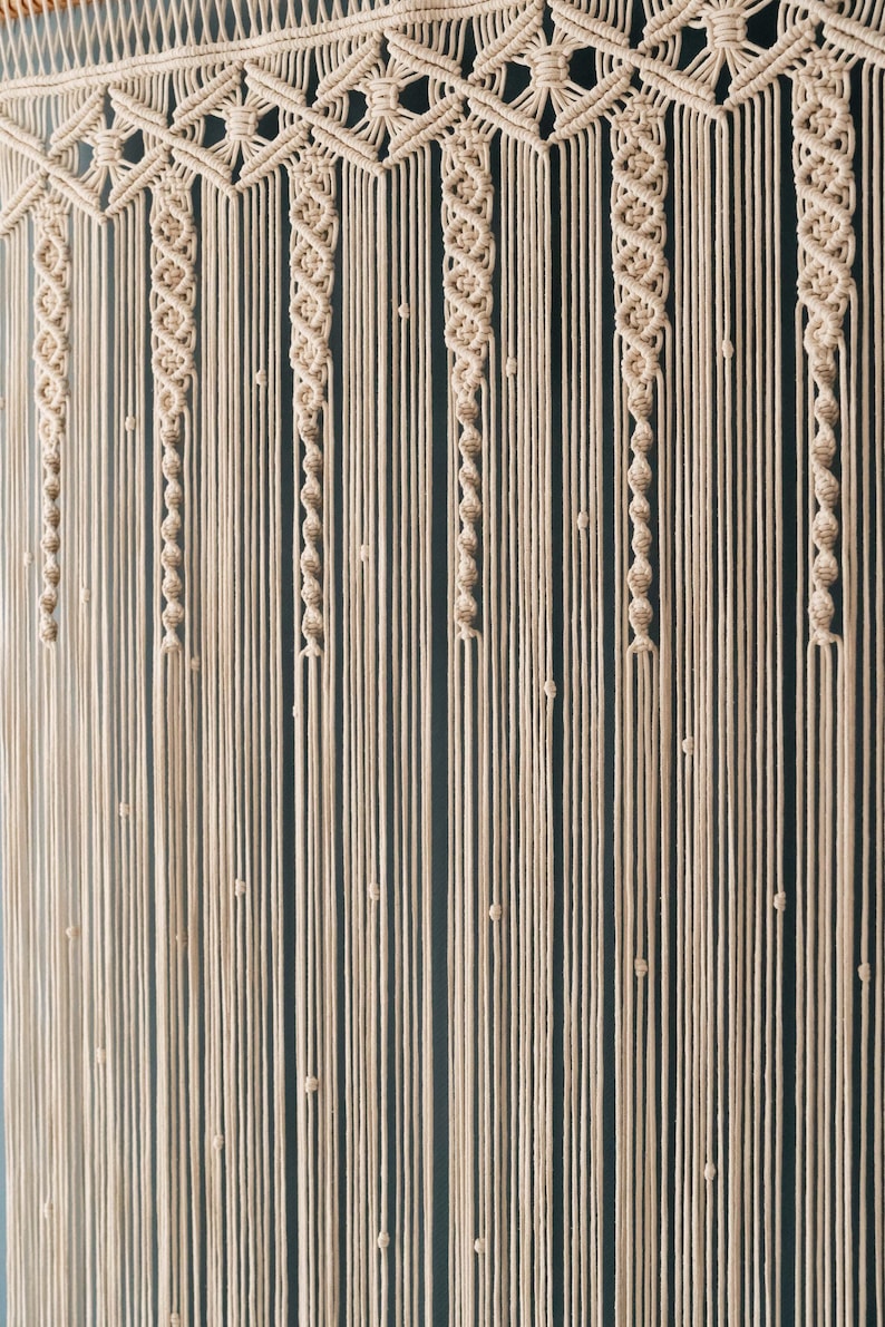 K&ouml;nnte beinhalten: Ein Makramee-Wandbehang mit geometrischem Muster und nat&uuml;rlicher beige Farbe. Der Behang ist aus dickem Baumwollschnur gefertigt und hat unten einen Fransen.