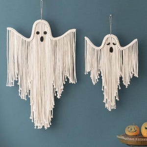 May include: Two off-white macrame ghost wall hangings made of knotted cord. The larger ghost features round black felt eyes and an oval mouth, while the smaller ghost has arched black felt eyebrows and an oval mouth. Both hang from thin strings.