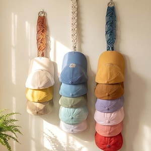 Könnte beinhalten: Drei Makramee-Huthalter mit Baseballkappen in verschiedenen Farben hängen an einer Wand. Die Halter sind in Orange-, Creme- und Blautönen gehalten. Die Kappen umfassen Weiß, Gelb, Beige, Blau, Grün und Rosa.