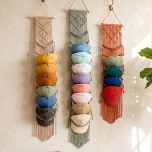 Può includere: Tre organizzatori di cappelli in macramè beige, verde salvia e arancione sono appesi a una parete. Ogni organizzatore contiene una serie di cappellini da baseball in vari colori, tra cui giallo, blu e rosso. Gli organizzatori hanno un design intrecciato con frange.