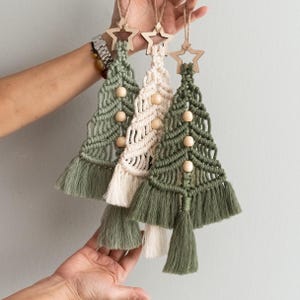 Può includere: Tre decorazioni per l'albero di Natale in macramè fatte a mano con stelle in legno. Le decorazioni sono in tonalità di verde e crema con perline di legno.