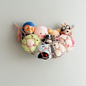 Op de afbeelding: Een roze en witte hangmat met een verscheidenheid aan pluche speelgoed erin, waaronder een pingu&iuml;n, een wasbeer, een kat en een kikker. De hangmat hangt aan een muur.