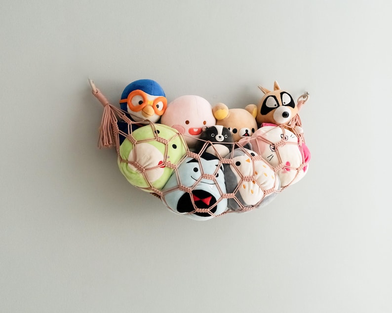 Op de afbeelding: Een roze en witte hangmat met een verscheidenheid aan pluche speelgoed erin, waaronder een pingu&iuml;n, een wasbeer, een kat en een kikker. De hangmat hangt aan een muur.