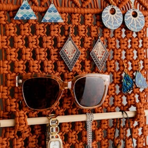 May include: A macrame wall hanging with a wooden dowel rod, featuring a variety of jewellery, including earrings, sunglasses, a watch, and necklaces. The wall hanging is made with brown cord and has a fringe at the bottom.