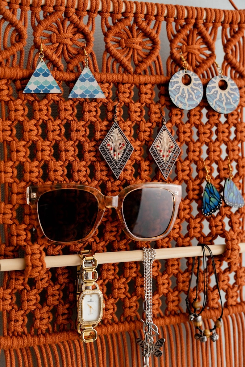 May include: A macrame wall hanging with a wooden dowel rod, featuring a variety of jewellery, including earrings, sunglasses, a watch, and necklaces. The wall hanging is made with brown cord and has a fringe at the bottom.