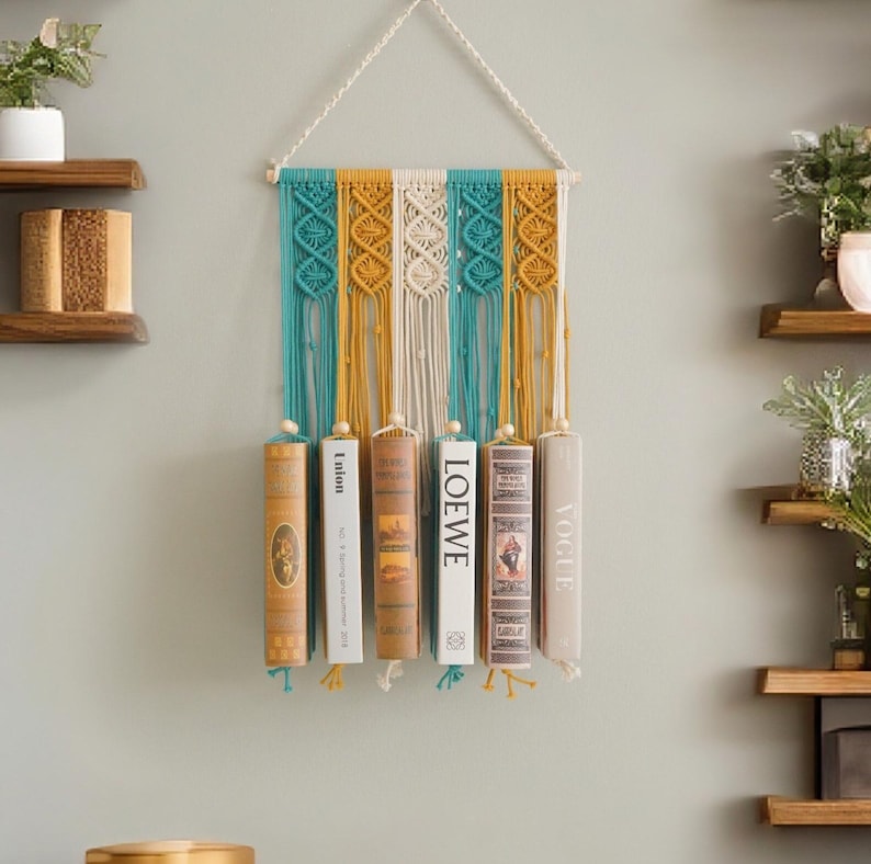 May include: A macrame wall hanging with a teal, white and mustard yellow colour scheme. The hanging has six wooden dowels that hold books in place.