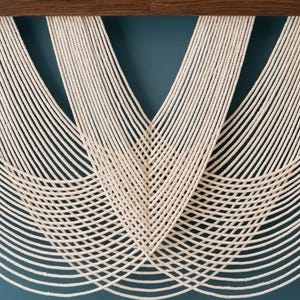May include: A wall hanging featuring multiple strands of off-white cotton rope suspended from a horizontal dark brown wooden bar. The ropes are arranged in overlapping, symmetrical U-shaped curves that create a layered, rhythmic pattern.