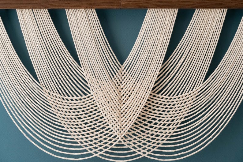 May include: A wall hanging featuring multiple strands of off-white cotton rope suspended from a horizontal dark brown wooden bar. The ropes are arranged in overlapping, symmetrical U-shaped curves that create a layered, rhythmic pattern.