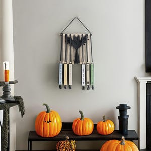 May include: A macrame wall hanging with a black and white geometric design. Five books are displayed on the wall hanging, with titles including "Book of Blood", "After the Fall", "Atlas of the Heart", and "Butter".  The wall hanging is displayed against a white wall with pumpkins in the foreground.