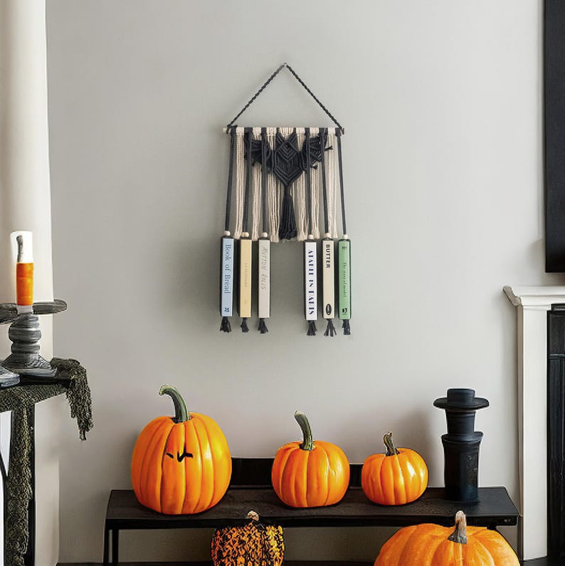 May include: A macrame wall hanging with a black and white geometric design. Five books are displayed on the wall hanging, with titles including "Book of Blood", "After the Fall", "Atlas of the Heart", and "Butter".  The wall hanging is displayed against a white wall with pumpkins in the foreground.