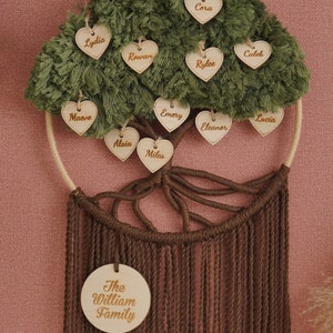 May include: A green and brown family tree wall hanging with wooden hearts featuring names like Cora, Rowan, Rylee, Caleb, Lucia, Emery, Eleanor, Maeve, Alaia, and Miles. The bottom of the hanging features a round wooden sign with the text "The William Family".