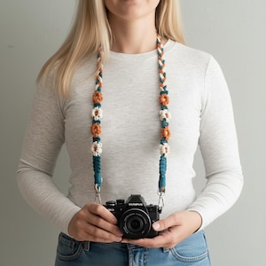 May include: A macrame camera strap with a braided design in shades of teal, orange, and cream. The strap features decorative flower accents and is attached to a black Olympus camera. The camera is held by a person wearing a long-sleeved shirt and jeans.
