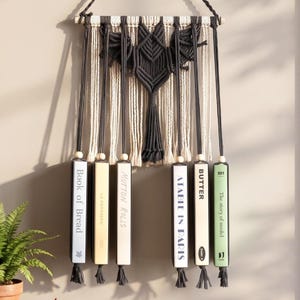 May include: A wall hanging featuring a black macrame bat centered above six vertical book-shaped blocks suspended by cords. The blocks display titles including Book of Bread, La Patisserie, Mutton Rolls, A Table in Paris, Butter, and The story of model 501.