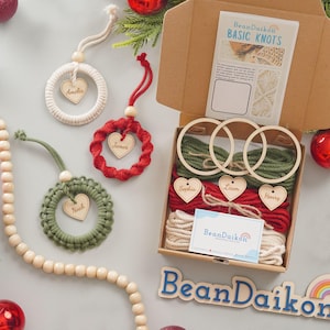 Puede incluir: Un kit de manualidades para hacer adornos navideños personalizados. El kit incluye tres aros de madera, hilo de color rojo, blanco y verde, y etiquetas de madera en forma de corazón con los nombres Amelia, James, Noah, Sophia, Liam y Henry. El kit está empaquetado en una caja marrón con las palabras "BeanDaikon BASIC KNOTS" impresas en la parte superior.