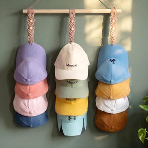 Peut inclure: Un porte-chapeaux en bois avec des suspensions en macramé présente une collection de casquettes de baseball de différentes couleurs. Les casquettes comprennent des nuances de bleu, de rose, de jaune et de marron. Certaines casquettes portent le mot "Memorie", d'autres des logos.