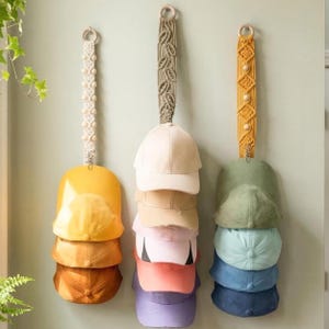 Könnte beinhalten: Drei Hut-Organizer aus Makramee hängen an einer Wand und halten jeweils einen Stapel Baseballkappen. Die Kappen sind in verschiedenen Farben erhältlich, darunter Gelb, Beige, Rosa und Blau. Die Makramee-Aufhänger haben Holzringe und dekorative Muster.