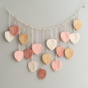 K&ouml;nnte beinhalten: Eine Girlande aus rosa, beige und wei&szlig;en Garnquasten in Form von Bl&auml;ttern. Die Quasten sind auf einer wei&szlig;en Schnur mit Holzperlen aufgef&auml;delt.
