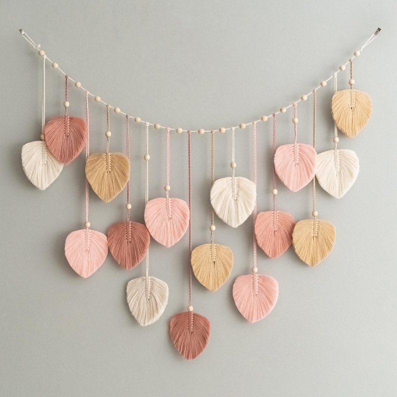 K&ouml;nnte beinhalten: Eine Girlande aus rosa, beige und wei&szlig;en Garnquasten in Form von Bl&auml;ttern. Die Quasten sind auf einer wei&szlig;en Schnur mit Holzperlen aufgef&auml;delt.