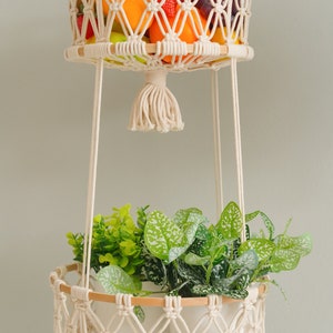 May include: Two hanging macrame plant hangers with wooden rings. The top hanger is filled with fruit, including oranges, apples, and grapes. The bottom hanger has a white pot with green leafy plants.
