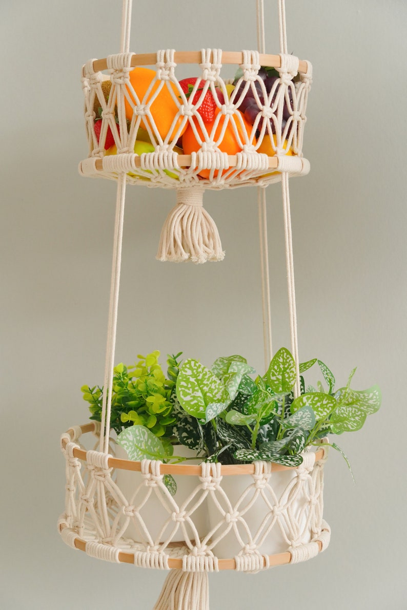 May include: Two hanging macrame plant hangers with wooden rings. The top hanger is filled with fruit, including oranges, apples, and grapes. The bottom hanger has a white pot with green leafy plants.
