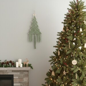 May include: A sage green macrame wall hanging shaped like a Christmas tree, featuring a star at the top, small wooden beads along the edges, and a long fringe at the bottom. The piece is constructed with intricate knotted cord patterns.