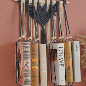 May include: A macrame wall hanging with a bat design in dark gray and cream yarn. The hanging has wooden dowels at the top and bottom and is holding up several books with titles like "Vogue" and "Loewe".