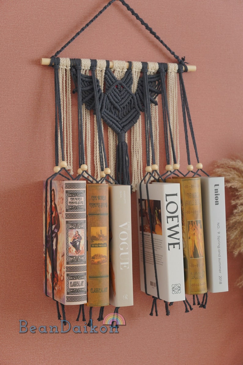 May include: A macrame wall hanging with a bat design in dark gray and cream yarn. The hanging has wooden dowels at the top and bottom and is holding up several books with titles like "Vogue" and "Loewe".