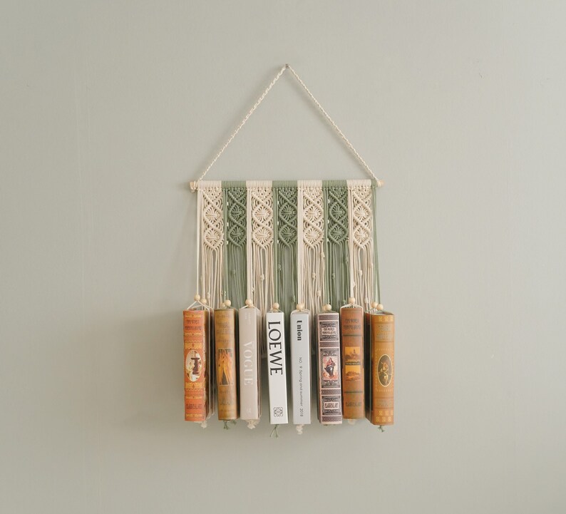 May include: A macrame wall hanging with a green and white pattern holds a row of books. The books have titles like "LOEWE" and "Vogue".