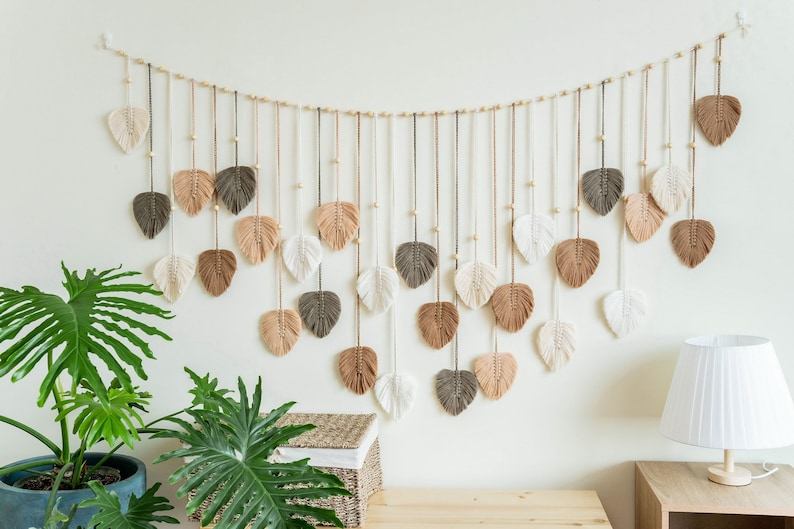 K&ouml;nnte beinhalten: Ein Wandbehang aus Makramee mit einer Reihe von braunen, beigefarbenen und wei&szlig;en Federformen. Der Behang ist an einer Holzleiste befestigt und hat kleine Holzperlen.