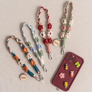 May include: A selection of handmade phone lanyards and a maroon phone case. The lanyards feature braided and macrame designs with flower accents in various colours, including orange, blue, and red. Each lanyard has a silver clasp and a wooden tag with a name.