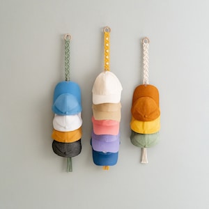 May include: Three macrame hat organizers hanging on a light gray wall. Each organizer holds a stack of baseball caps in various colors, including blue, white, yellow, and brown. The organizers have wooden rings and decorative macrame straps.