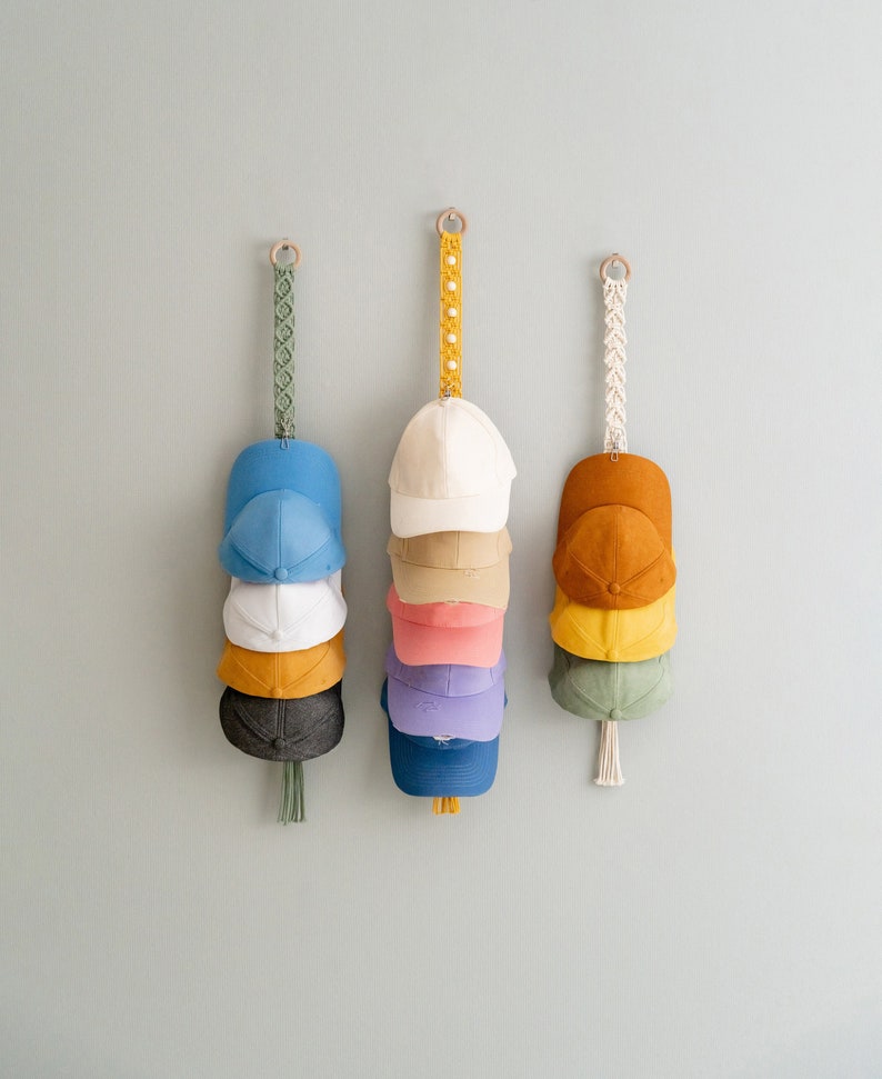 May include: Three macrame hat organizers hanging on a light gray wall. Each organizer holds a stack of baseball caps in various colors, including blue, white, yellow, and brown. The organizers have wooden rings and decorative macrame straps.